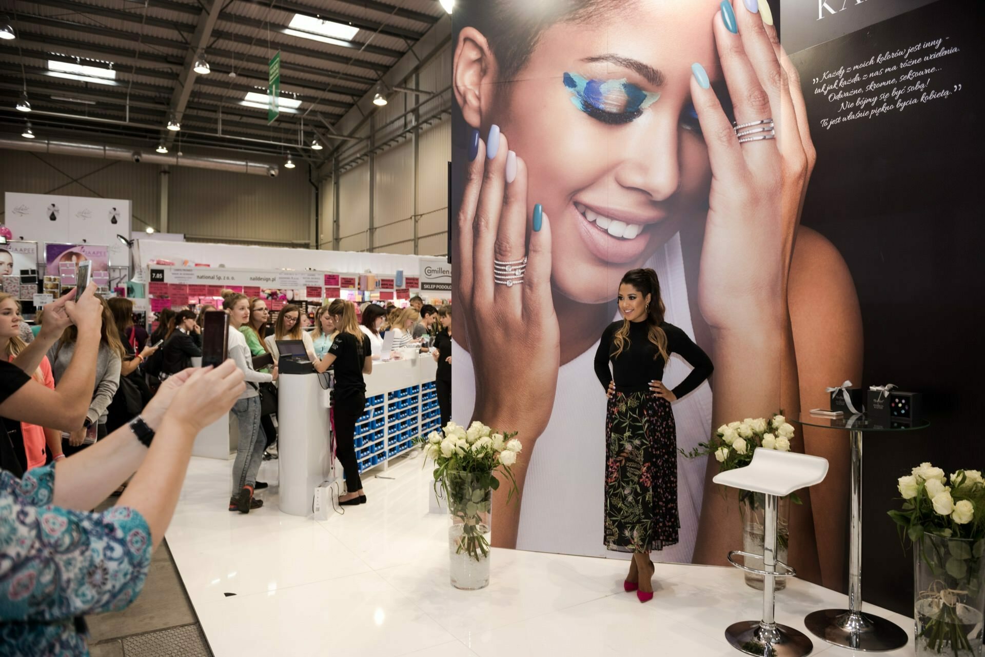 A woman poses for a photo in front of a large advertisement at a trade show or convention. She stands on a white floor decorated with stools and flower arrangements. Attendees, including well-known event photographer Marcin Krokowski, capture her image against the backdrop of various booths and exhibits.  