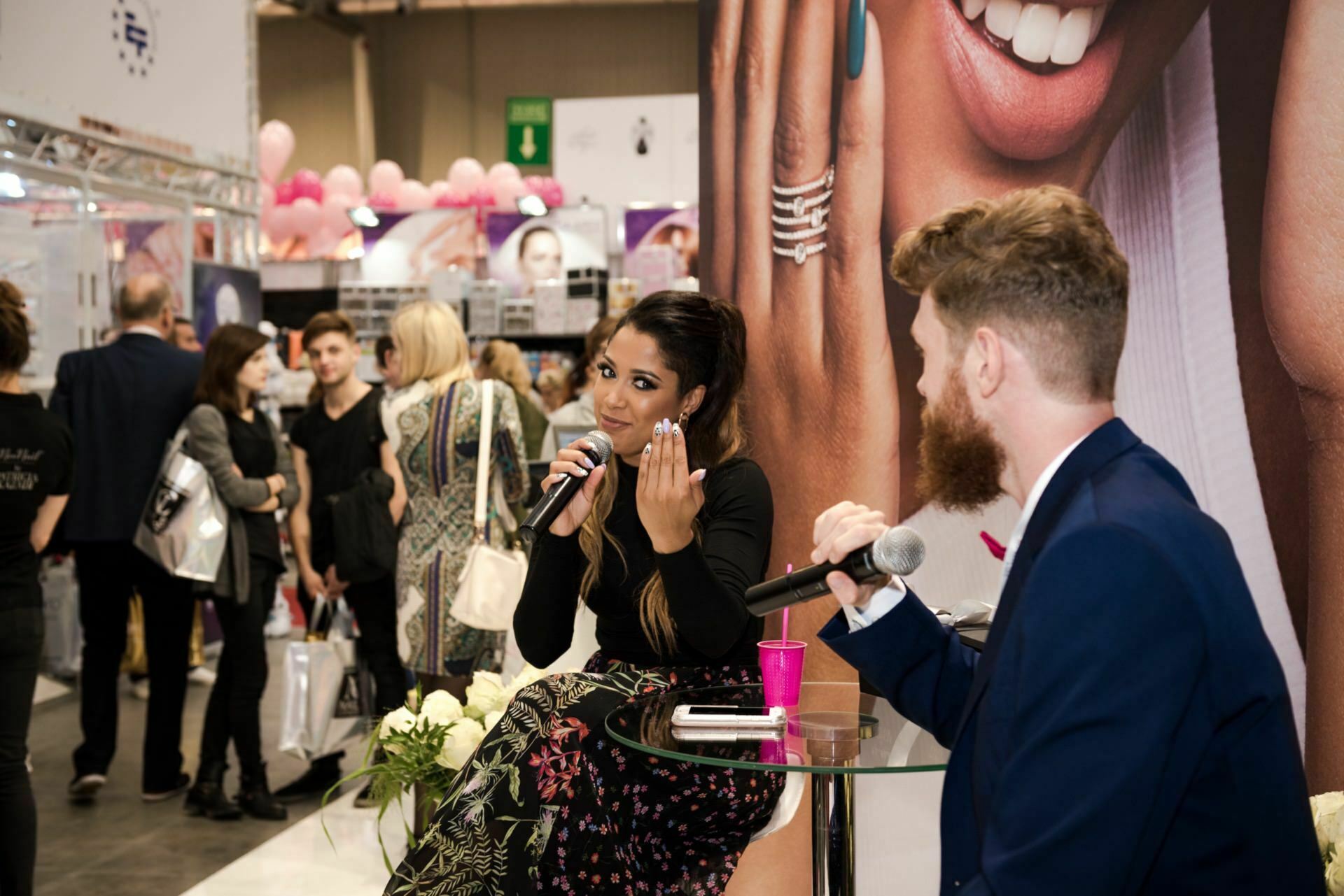 A woman and a man sit and talk into microphones in what resembles a bustling Beauty Forum. The woman gestures with her hand, the man listens intently. A crowd can be seen in the background, followed by a large advertising banner.  