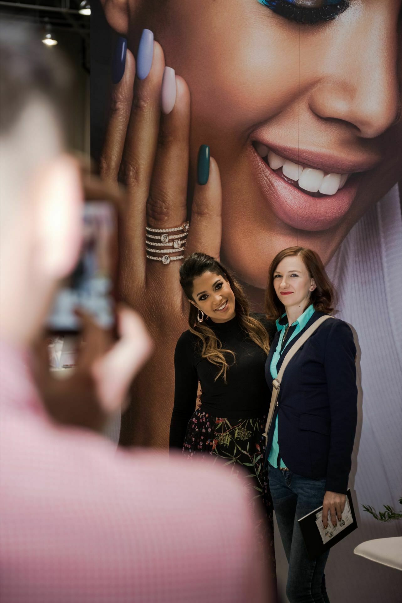 Two women pose for a photo in front of a large advertisement, which shows a close-up of the woman's smiling face and colorful, manicured nails. The photo was taken by Marcin Krokowski, a well-known photographer in Warsaw, who captured the couple smiling in front of the vibrant poster. 