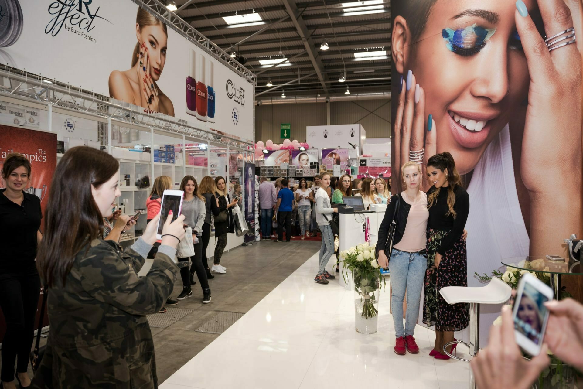 The bustling convention center showcases various booths of beauty products. People are hanging around, some taking pictures. One booth is decorated with a large poster showing a smiling woman with colorful eye makeup. Event photographer Marcin Krokowski captured two women posing for a photo, taken by another in the foreground.   