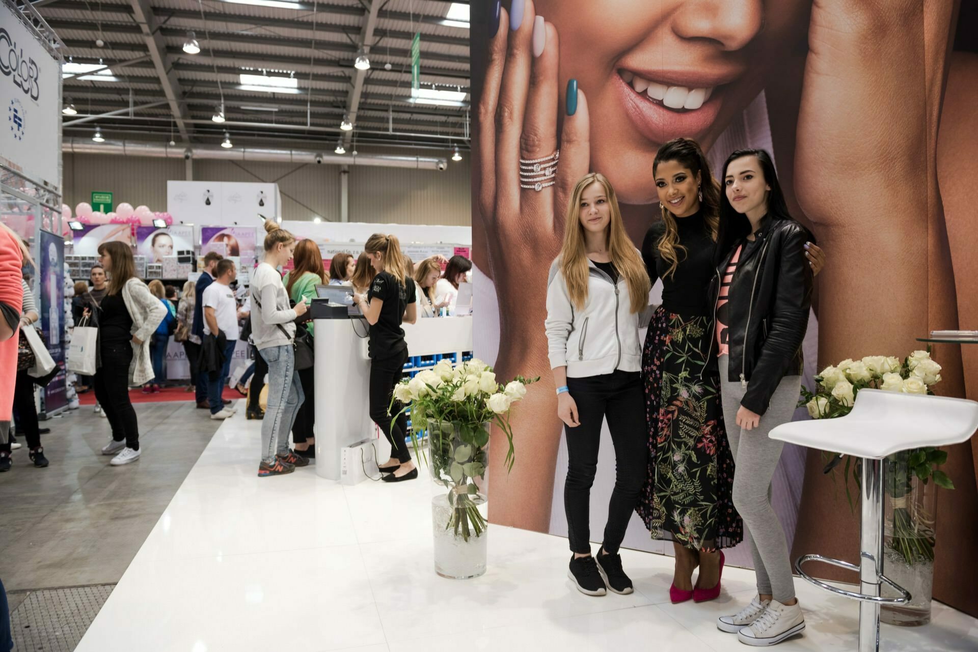 Three women stand together in front of a large cosmetics advertisement at a trade show, captured by talented event photographer Marcin Krokowski. They smile at the camera. The booth is decorated with white roses. In the background, attendees visit other booths in a spacious, well-lit room.   