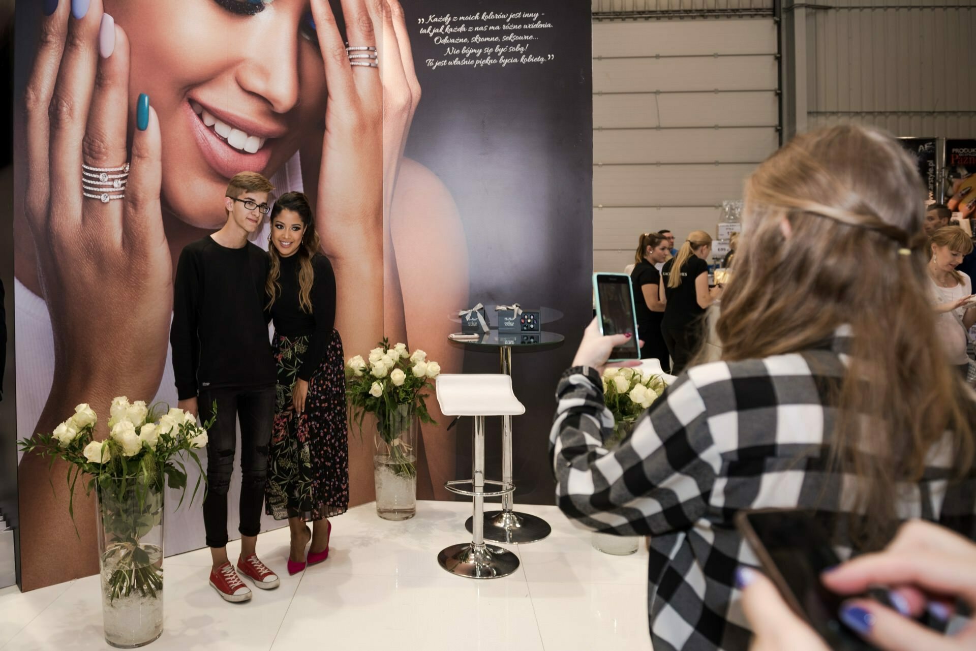 At a jewelry event, a young man and woman pose for a photo. Behind them, a banner shows a woman adorned with rings. White flowers decorate the area, while another person in a checked shirt captures the moment on his smartphone. Marcin Krokowski, a Warsaw-based event photographer, captured the scene perfectly.   