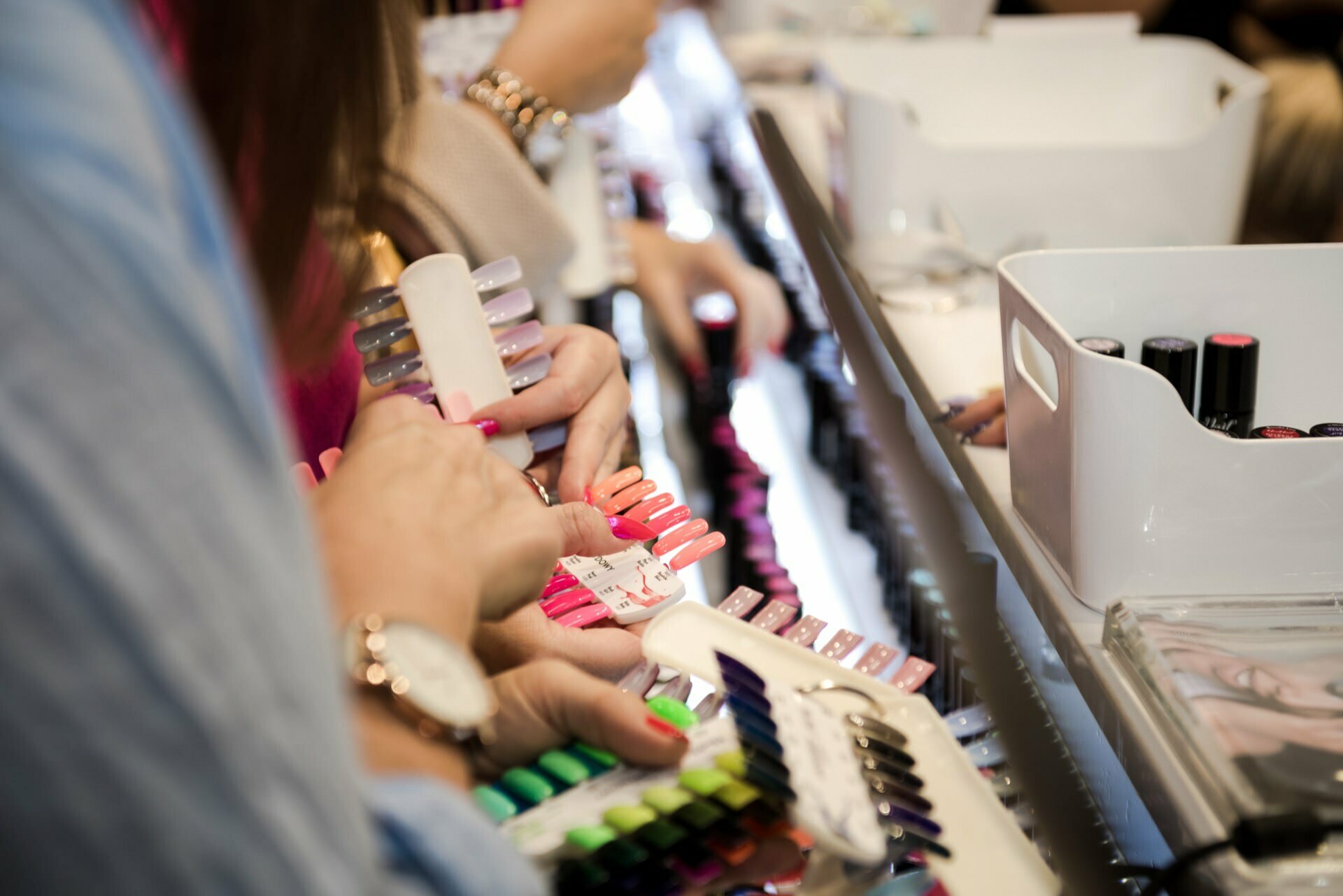 Close-up of several people choosing nail polish colors from sample palettes at a nail salon. They are holding various color samples and looking at the different shades. Nail polish bottles and cosmetic utensils are visible in the background, beautifully captured by Marcin Krokowski, a talented event photographer in Warsaw.  