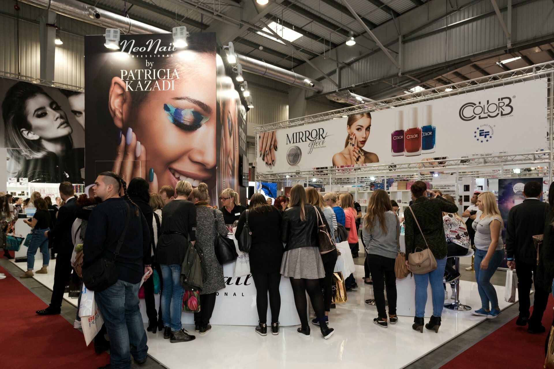 The crowded beauty fair includes attendees gathered around various exhibitors' booths. Large banners display photos of models and brands such as NeoNail, Patricia Kazadi, Mirror Glass and Color that Lasts. The venue is spacious, with industrial metal beams, perfectly captured by event photographer Marcin Krokowski.  