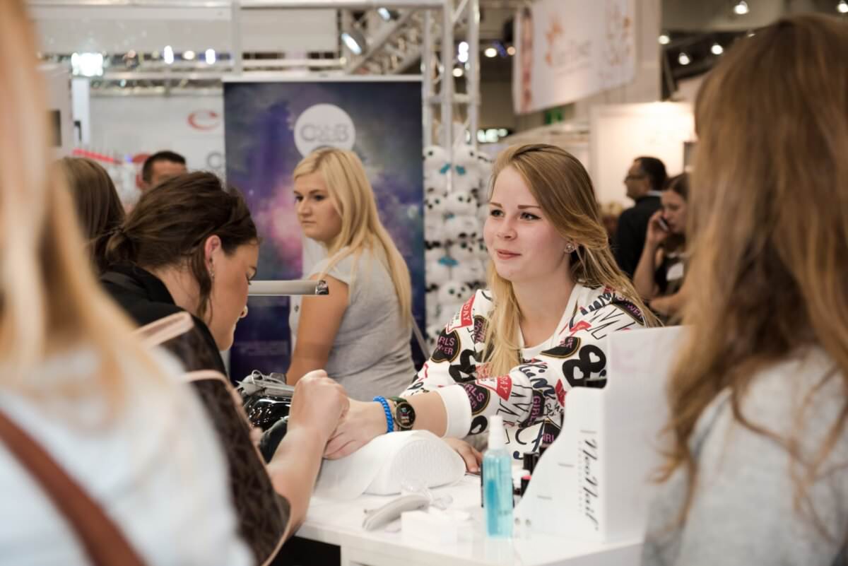 Kobieta ubrana w białą bluzę z kolorowymi wzorami otrzymuje manicure przy stole podczas pracowitego wydarzenia. Uchwycone przez Marcina Krokowskiego, fotografa eventowego w Warszawie, w tle widać innych uczestników i stoiska, co podkreśla żywą i interaktywną atmosferę.