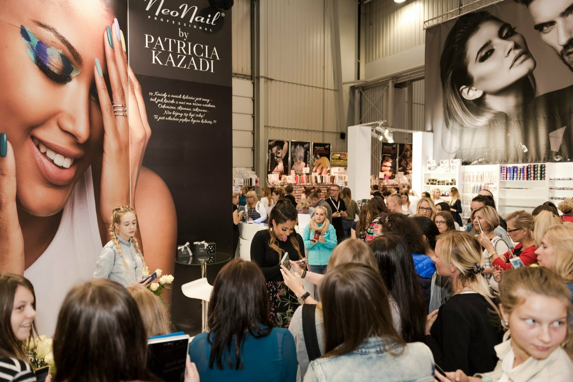 A diverse crowd gathers around a booth in a busy exhibition, on which hangs a large poster depicting a smiling woman with colorful eye makeup, advertising NeoNail by Patricia Kazadi. Marcin Krokowski, a well-known Warsaw event photographer, captures the participants engaging and taking photos of themselves with various products displayed in the background. 