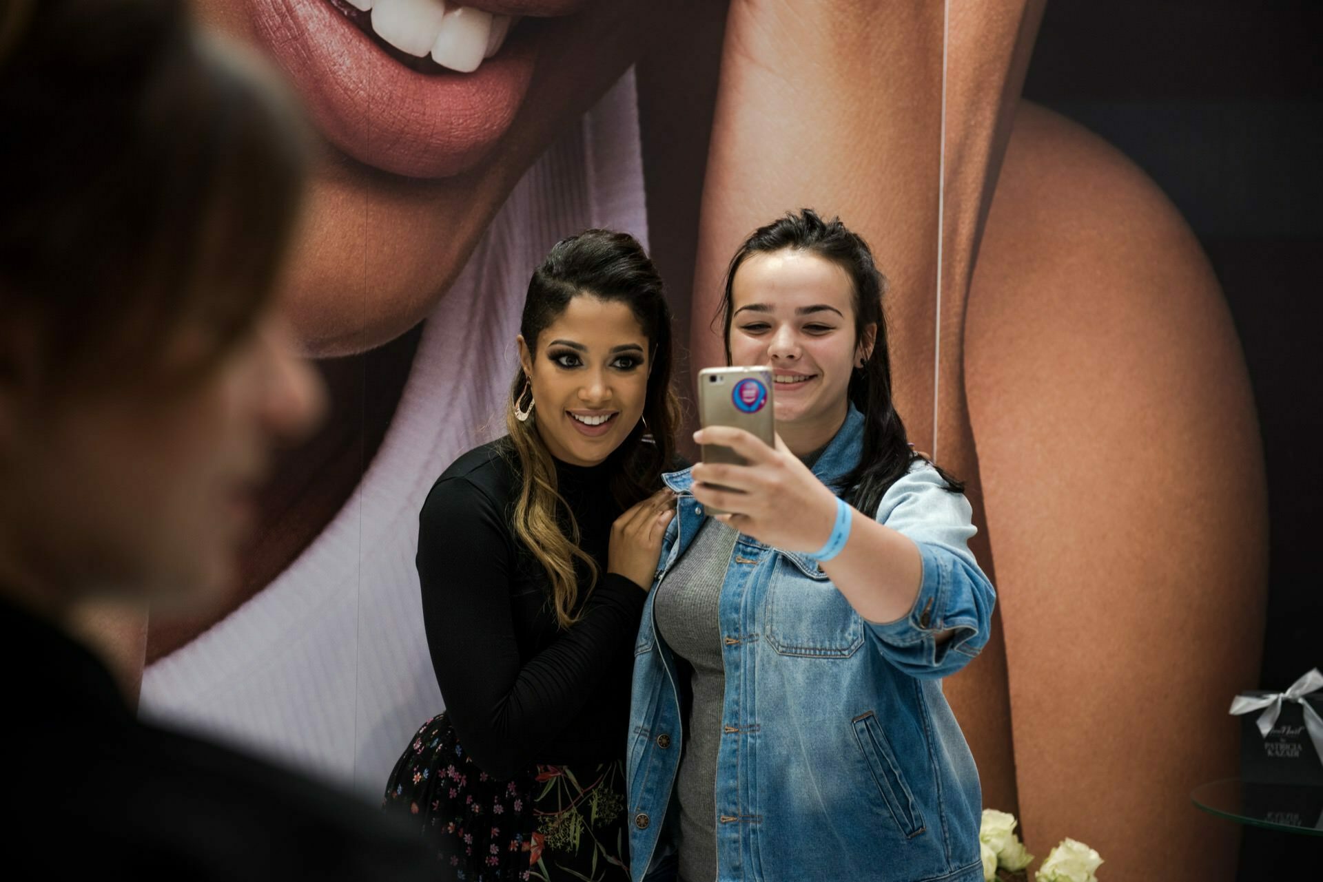 Two women take a selfie together. The woman on the left has long, dark hair and is smiling, while the woman on the right is holding her phone and smiling. They look as if they are at some event in Warsaw, with a large poster depicting a smiling mouth in the background. A perfect shot for an event photographer like Marcin Krokowski.   