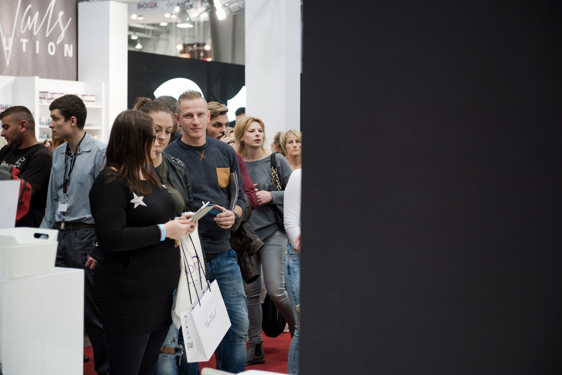 A group of people are standing in line at an event held indoors. A woman in front is holding a bag and some papers. Behind her, a man and several other people are looking ahead, and some are talking to each other. The area is well-lit, with exhibits and booths visible in the background, whose photos were taken by Marcin Krokowski, an established event photographer in Warsaw.   