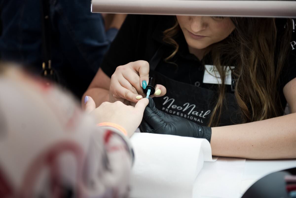 Zbliżenie na stylistkę paznokci nakładającą ciemnozielony lakier na paznokcie klientki. Technik ubrany jest w czarny fartuch z napisem „NeoNail Professional” i czarne rękawiczki. Dłoń klienta spoczywa na białym ręczniku. To zdjęcie, wykonane przez Marcina Krokowskiego, fotografa w Warszawie, ukazuje szczegółowe rzemiosło.