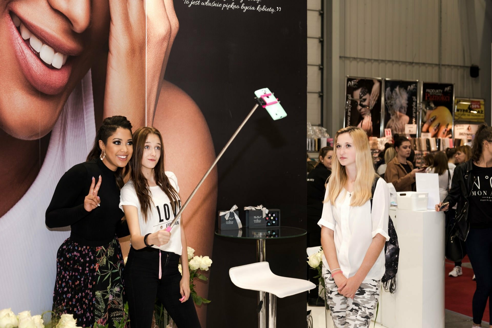 Two women take a selfie using a selfie stick at an event with beauty posters in the background. One woman makes a peace sign, while the other holds the selfie stick. A third woman, watching the event, seems intrigued by the fun captured by Marcin Krokowski, a well-known event photographer in Warsaw.  