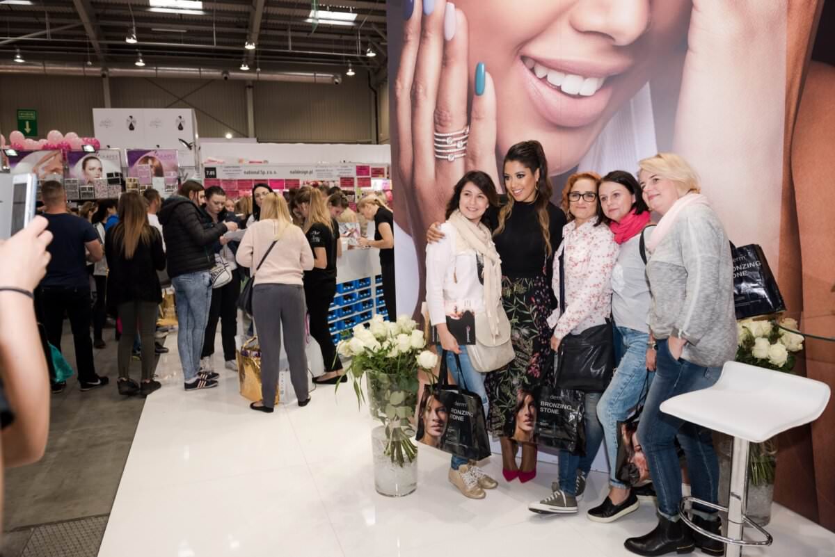 Grupa czterech kobiet pozuje do zdjęcia wykonanego przez znanego fotografa eventowego Marcina Krokowskiego przed dużym plakatem przedstawiającym uśmiechniętą kobietę z wypielęgnowanymi dłońmi podczas ruchliwej imprezy. Inni uczestnicy przeglądają stoiska w tle. Kobiety są ubrane zwyczajnie i trzymają torby na zakupy, na których widać twarz modelki.