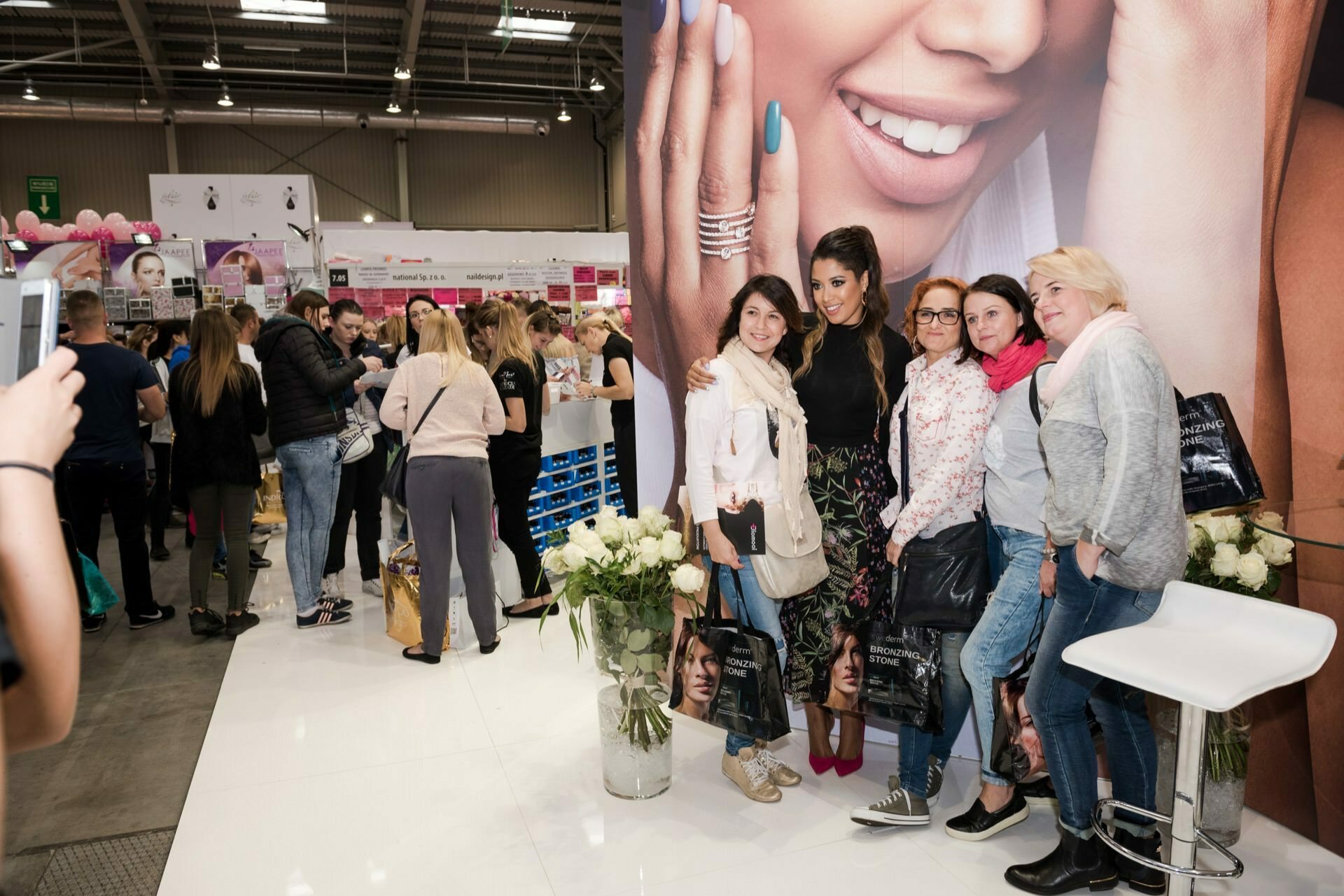 A group of four women pose for a photo taken by well-known event photographer Marcin Krokowski in front of a large poster showing a smiling woman with manicured hands during a busy event. Other attendees browse the booths in the background. The women are dressed casually and holding shopping bags that show the model's face.  