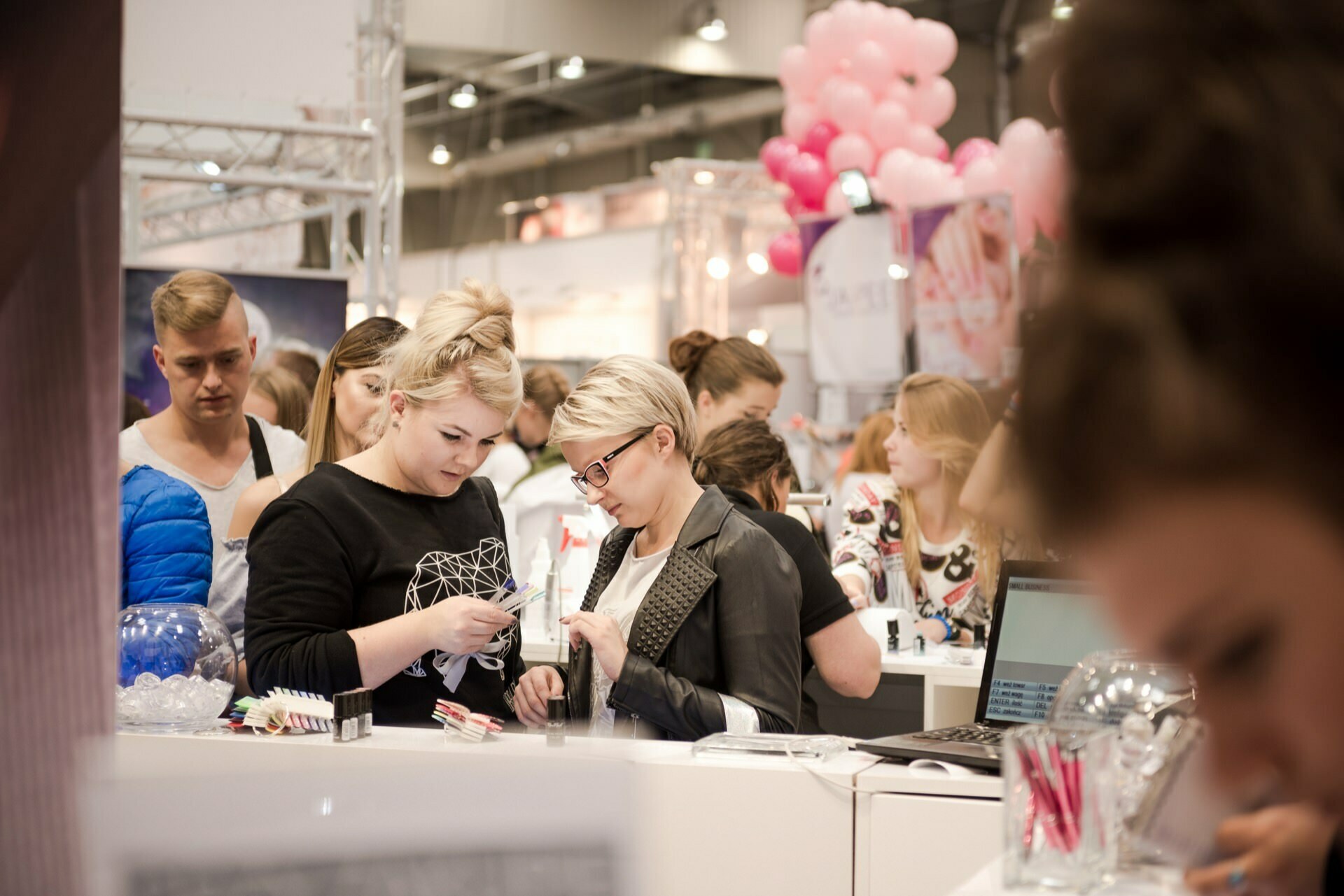 A busy exhibition hall showing a crowd of people interacting at a booth. Two women in the foreground are looking at products with various items and a laptop on the tabletop. In the background you can see other attendees, balloons and exhibition stands, all captured by Marcin Krokowski, an acclaimed event photographer.  