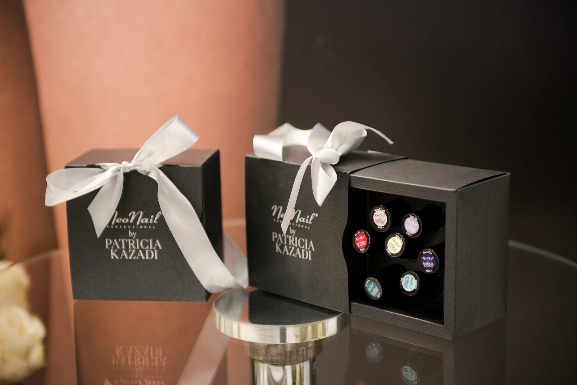 Two black gift boxes are placed on a reflective surface. Each box, tied with a white ribbon, contains nail polish bottles with different colored caps. The boxes feature the brand "NeoNail" and the name "Patricia Kazadi" written in white text, painstakingly captured by photographer in Warsaw, Marcin Krokowski.  