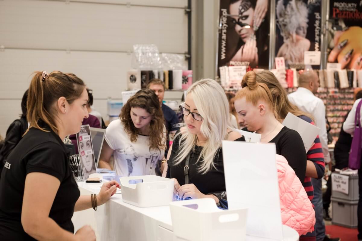 Grupa ludzi gromadzi się wokół lady podczas konwencji lub targów. Pracownica pomaga im, gdy uczestnicy trzymają torby i broszury. W tle eksponowane są rozmaite produkty kosmetyczne i plakaty, doskonale uchwycone przez znanego warszawskiego fotografa Marcina Krokowskiego.