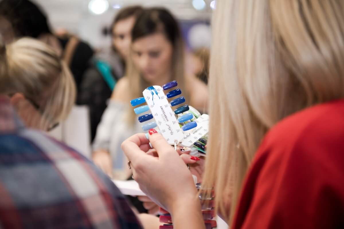 Osoba trzyma wystawę próbek wielokolorowego lakieru do paznokci w zatłoczonym otoczeniu, a w tle widać kilka innych osób. Scena ta, uchwycona przez fotografa eventowego Marcina Krokowskiego, doskonale oddaje tętniącą życiem atmosferę takich wydarzeń.