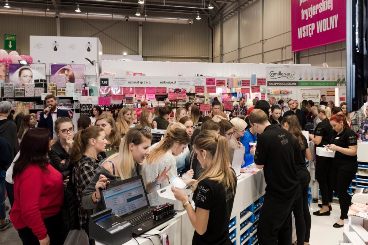 Na tętniącym życiem stoisku targów kosmetycznych tłumy chętnych uczestników wchodzą w interakcję z personelem. Kobiety oglądają produkty, biorą udział w demonstracjach i robią notatki. Stoisko jest dobrze oświetlone, ekspozycje, półki z produktami oraz oznakowanie w języku polskim. Fotograf eventowy doskonale oddaje tętniącą życiem atmosferę.