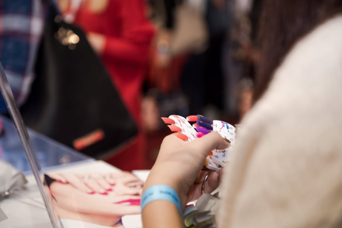 Osoba trzymająca zestaw próbek kolorów lakieru do paznokci na stanowisku do manicure. Osoba nosi niebieską opaskę, a w tle widać nieostre inne osoby, co tworzy ruchliwą atmosferę. Uchwycone przez Marcina Krokowskiego, fotografa eventowego w Warszawie.