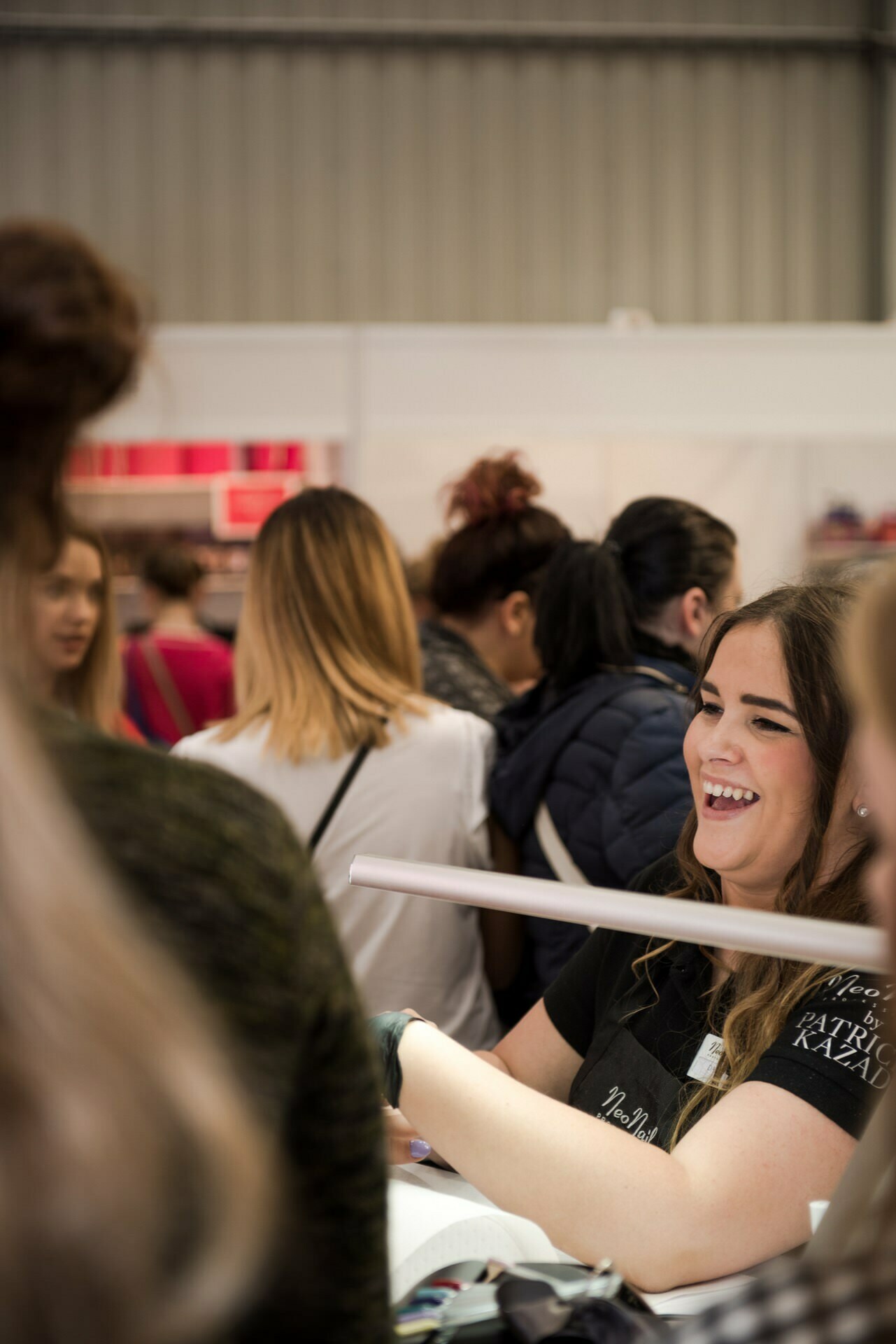 A woman with long brown hair, dressed in black, smiles and interacts with people at a crowded event or market. She appears to be signing something. Captured by photographer in Warsaw Marcin Krokowski, the background shows a mix of people with different hairstyles and outfits.  