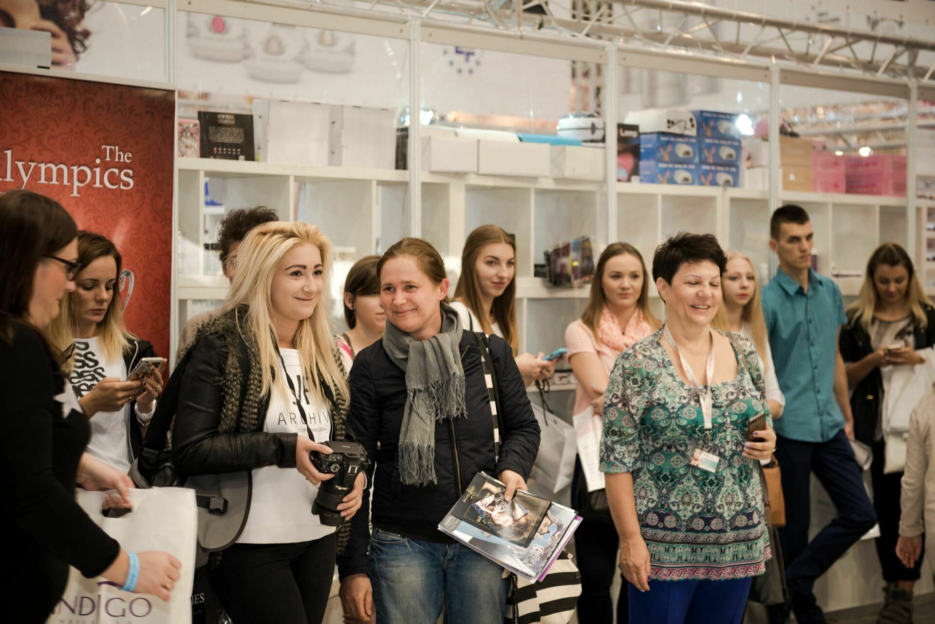 A group of people are standing in the Beauty Forum room. Some are smiling, while others are using their phones. Shelves of various products are visible in the background. One person is holding a camera and another is holding something that looks like a brochure or pamphlet.   