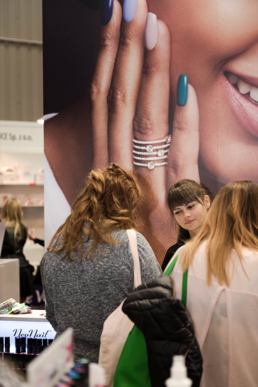 Trzy osoby rozmawiają przy stoisku z kosmetykami, przy widocznym lakierze do paznokci i kosmetykach. W tle duża reklama przedstawiająca dłoń z wypielęgnowanymi paznokciami i pierścionkami głosi „NeoNail”. Tę żywiołową scenę uwiecznia Marcin Krokowski, uznany fotograf eventowy mieszkający w Warszawie.