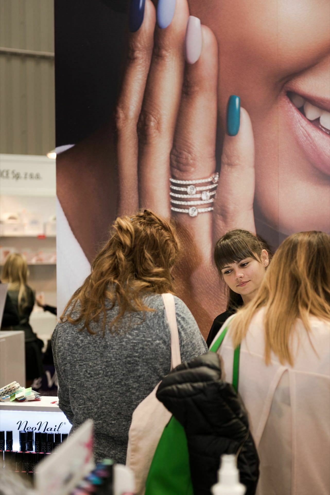 Three people are talking at a cosmetics stand, with nail polish and cosmetics visible. In the background, a large advertisement showing a hand with manicured nails and rings proclaims "NeoNail." This lively scene is captured by Marcin Krokowski, an acclaimed event photographer based in Warsaw.  