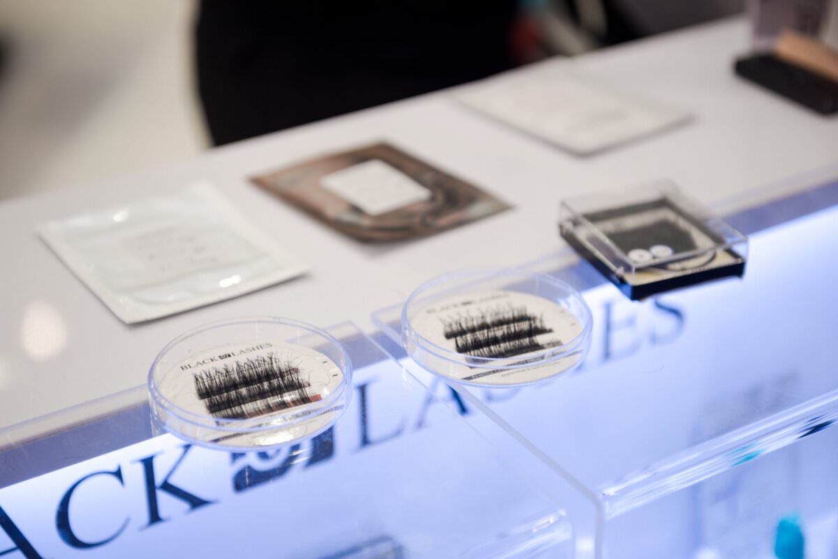 Display na ladzie prezentujący zapakowane przedłużacze do rzęs z przezroczystymi wieczkami, z etykietą „BLACK LASHES”, uchwycony przez Marcina Krokowskiego, wraz z innymi drobnymi przedmiotami i opakowaniami w tle, sugerującymi ustawienie sklepu z kosmetykami.