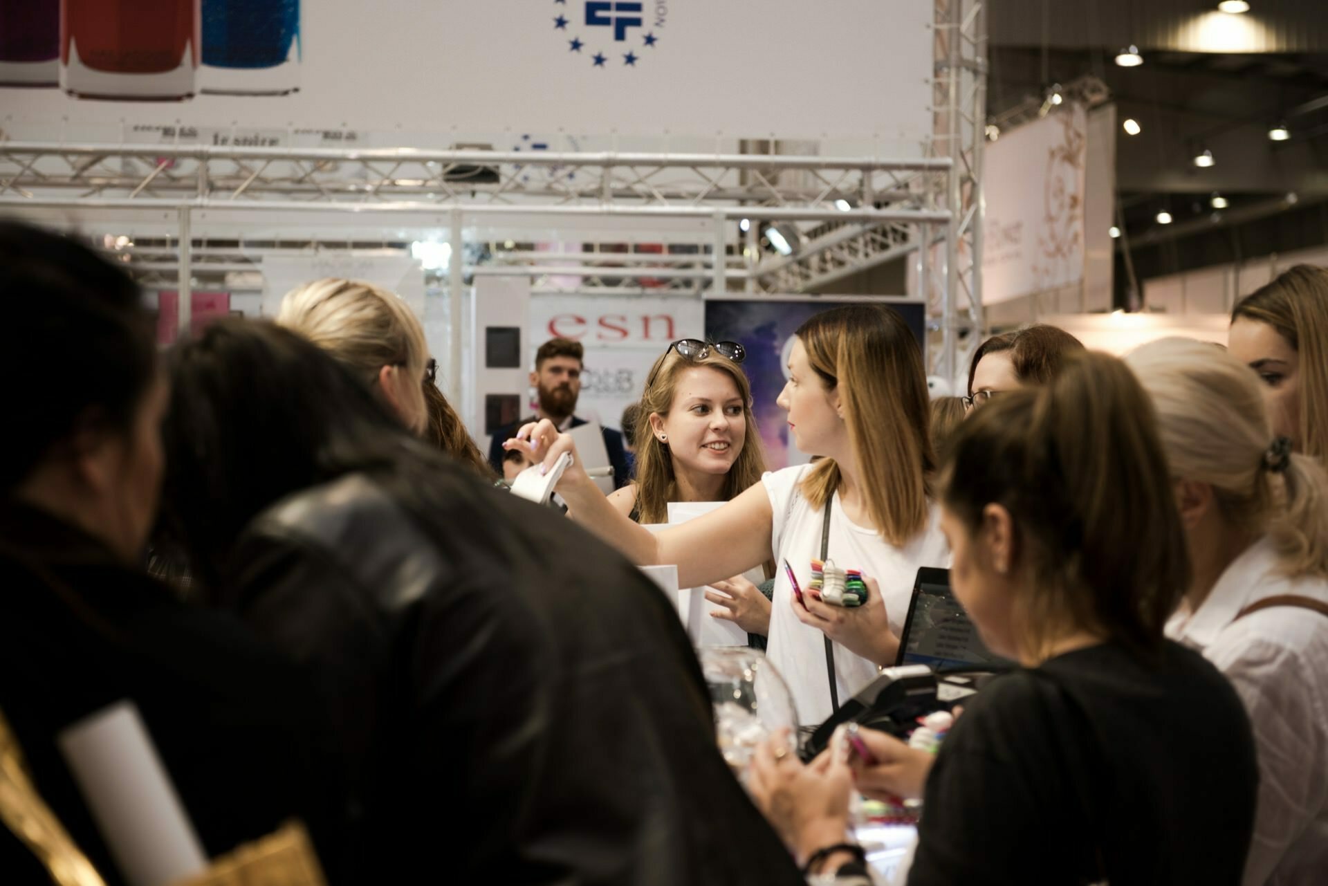 A group of people, mostly women, gather around a booth at a trade show or exhibition. They give the impression of being busy talking and browsing products. In the background are various banners and displays captured by Marcin Krokowski, a well-known Warsaw event photographer. The atmosphere seems busy and bustling.   