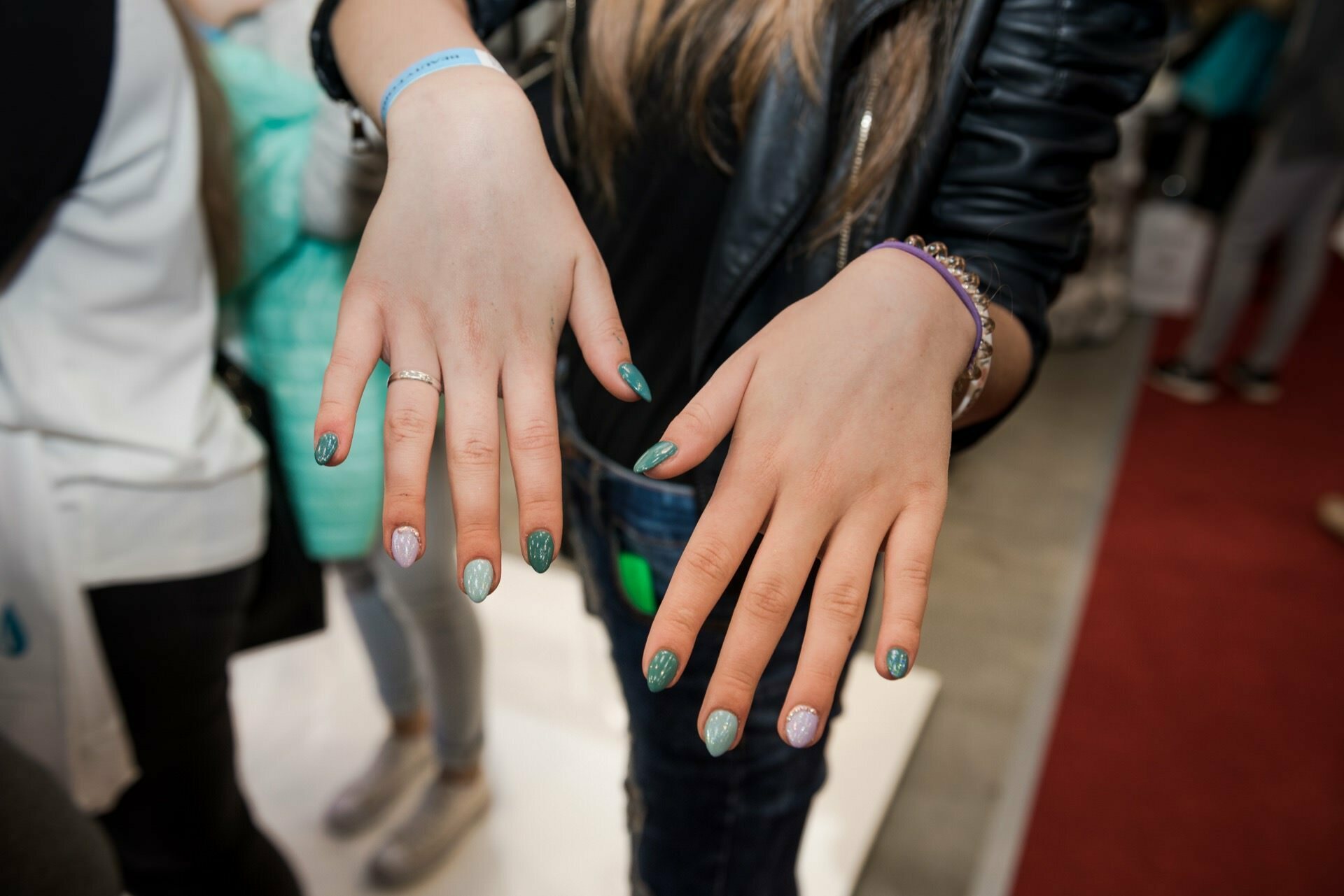 Close-up of a person's hands with freshly manicured nails. The nails are painted with glossy green polish, highlighted by intricate designs on the ring fingers. The person is wearing a black leather jacket and a colorful bracelet. Captured by Marcin Krokowski, a photographer in Warsaw, in a vibrant setting.   