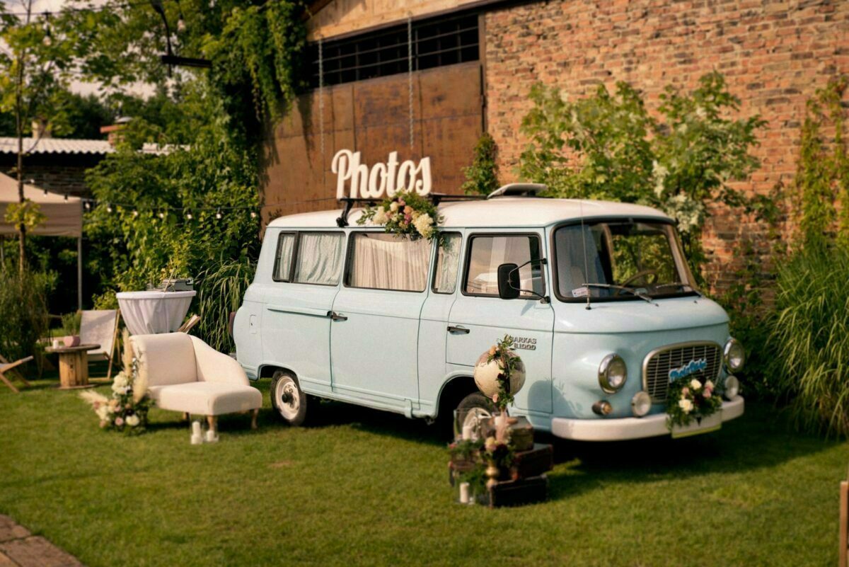 A light blue vintage-style van decorated with flowers stands parked on a grassy area near a brick wall with the word "Photos" on top. Nearby, a white couch, a small round table with a white tablecloth and assorted flower arrangements create a cozy outdoor setting, perfect for the shoot by Marcin Krokowski, a photographer in Warsaw. 