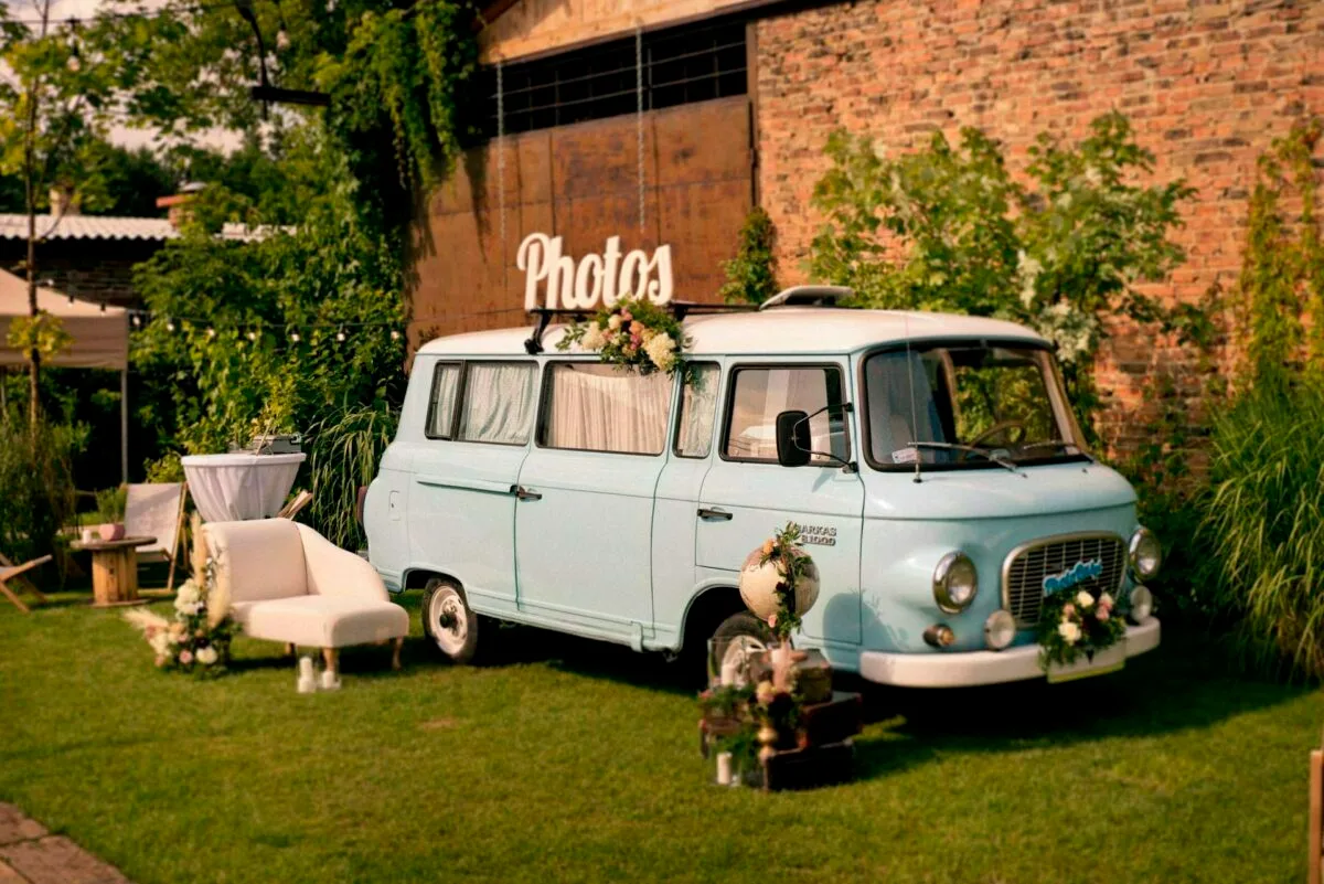 A light blue vintage-style van decorated with flowers stands parked on a grassy area near a brick wall with the word "Photos" on top. Nearby, a white couch, a small round table with a white tablecloth and assorted flower arrangements create a cozy outdoor setting, perfect for the shoot by Marcin Krokowski, a photographer in Warsaw. 