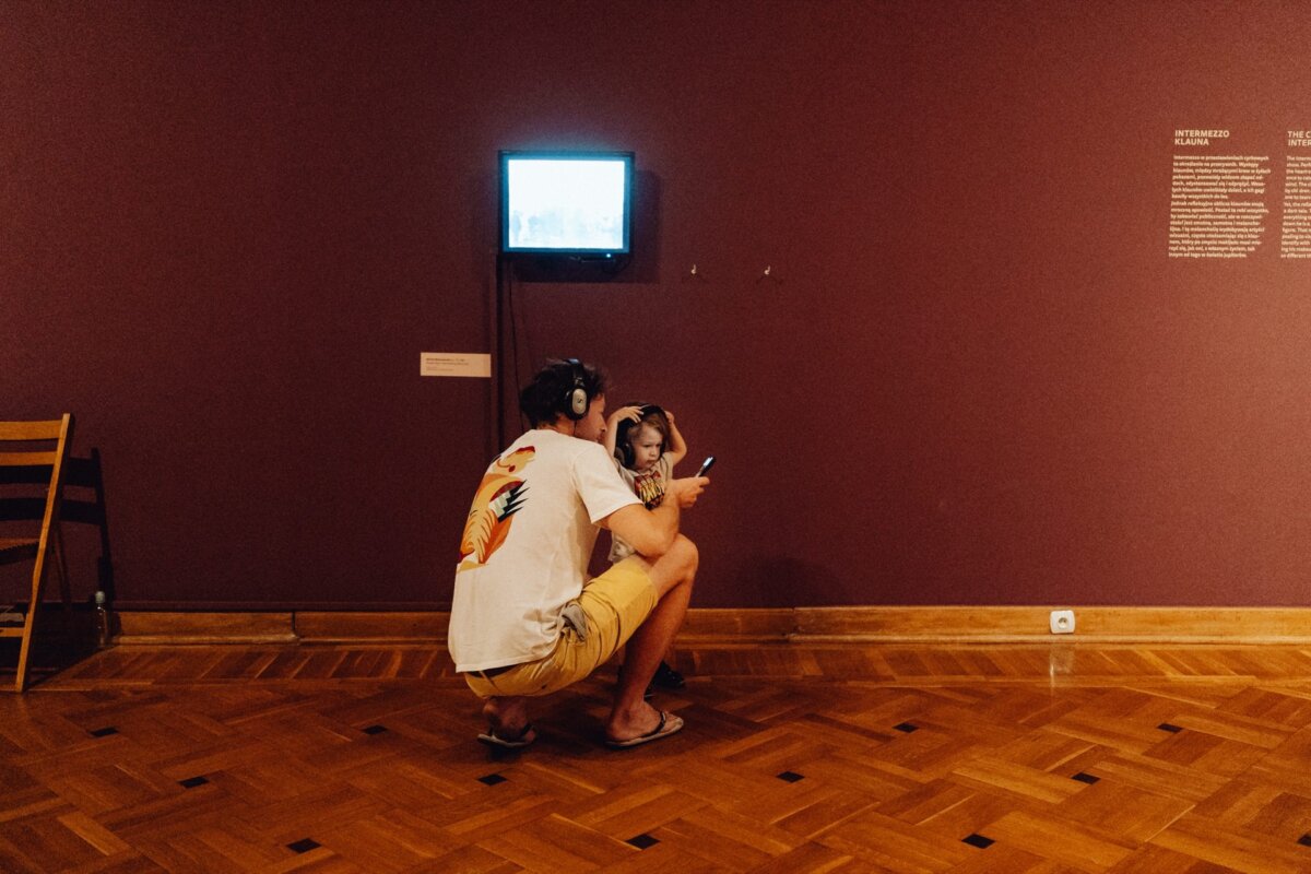 A man crouches next to a child in front of a small TV screen mounted on a purple wall in an art gallery. Both are wearing headphones and looking at the smartphone the child is holding. A wooden chair and a text board are visible in the background, perfectly captured by a photographer from Warsaw.  