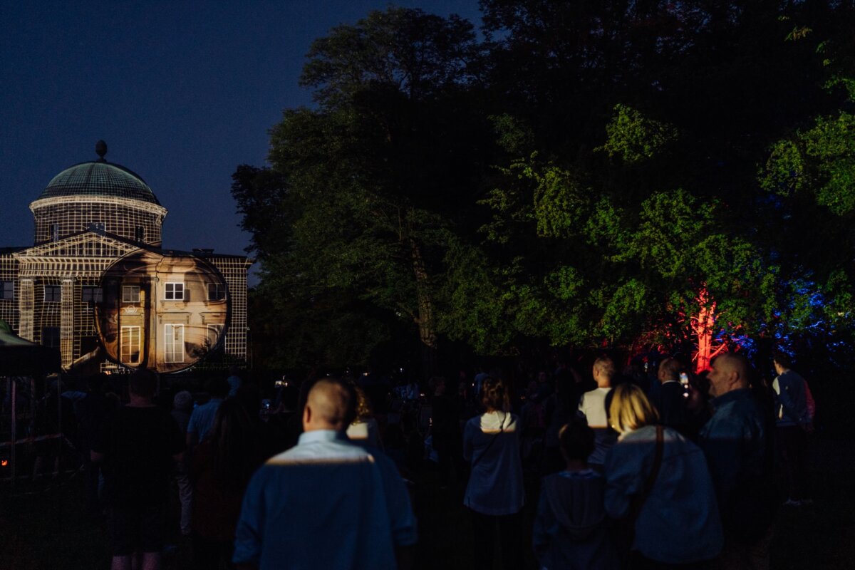 A nighttime outdoor event with people gathered in front of the building. The building has a projection of a detailed image of the house on the facade. The trees in the background are illuminated with vivid red and blue lights, which was perfectly captured by a photographer for the event from Warsaw.  