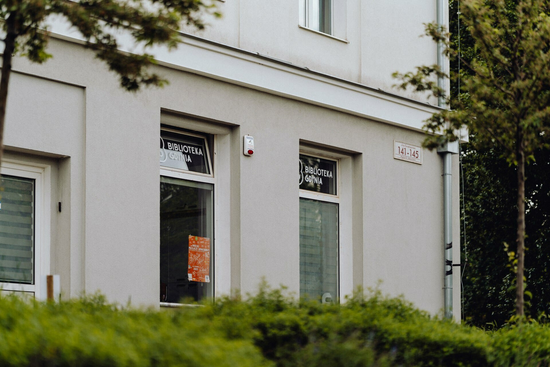 A gray two-story building with a sign in the window reading "Library." Trees and shrubs partially obscure the lower windows, adding to the scene's charm. The address "141-145" is visible outside. Perfect for a photographer from Warsaw to capture, this place has a peculiar charm.   