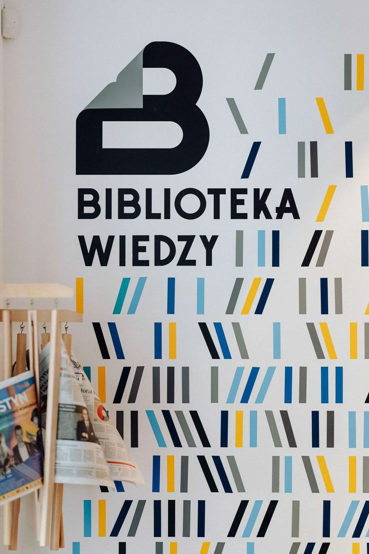 A wall with the logo and name "Knowledge Library" in Polish and a designed pattern of colorful lines. On the left side, a wooden gazetteer with several newspapers is visible - perfect for event photographer Warsaw to capture. 