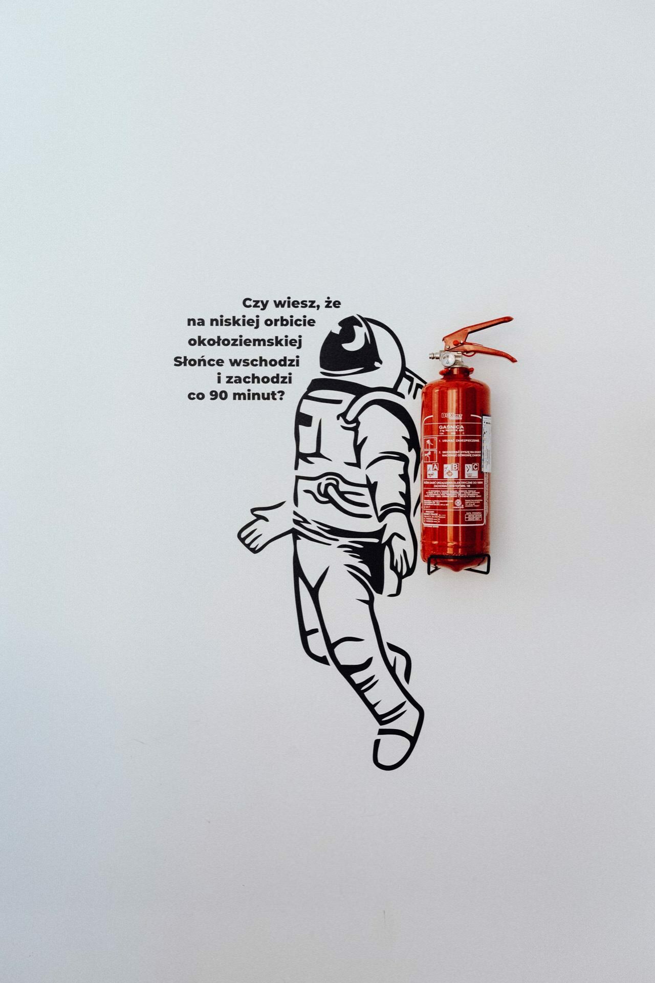 A mural depicting an astronaut with a red fire extinguisher embedded in the design, placed where the astronaut's backpack would stand. Above the astronaut the Polish text reads, "Did you know that on the orbital deck the sun rises and sets every 90 minutes?" This captivating piece would make a striking backdrop for a photo essay from the