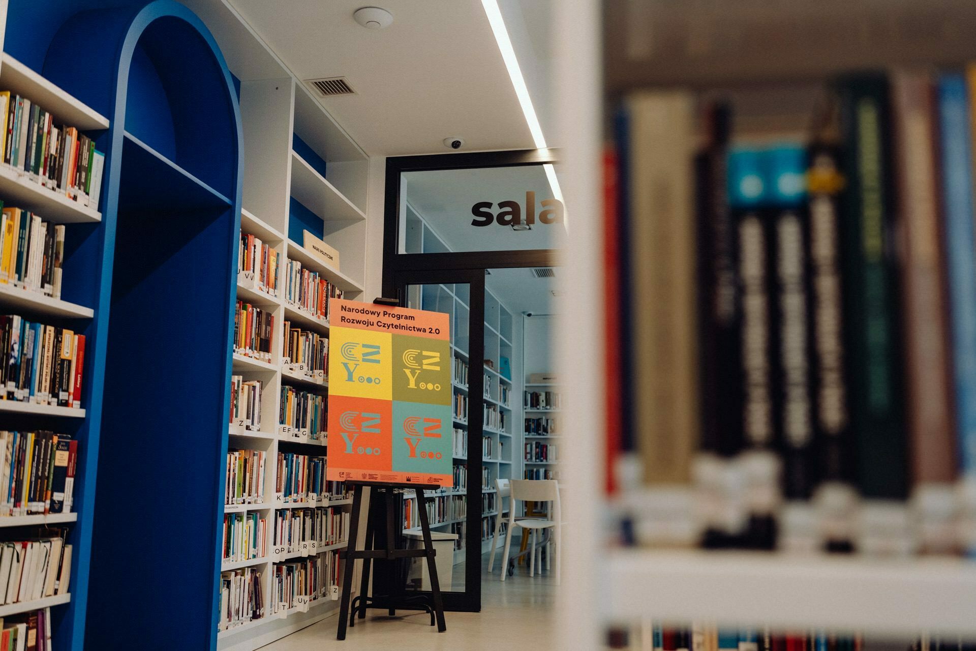 A modern library with white shelves filled with books on both sides. In the middle, a colorful poster on an easel promotes the event. Behind the poster, a glass door with the word "hall" leads to another part of the library, where you can see a photo essay of the event.  