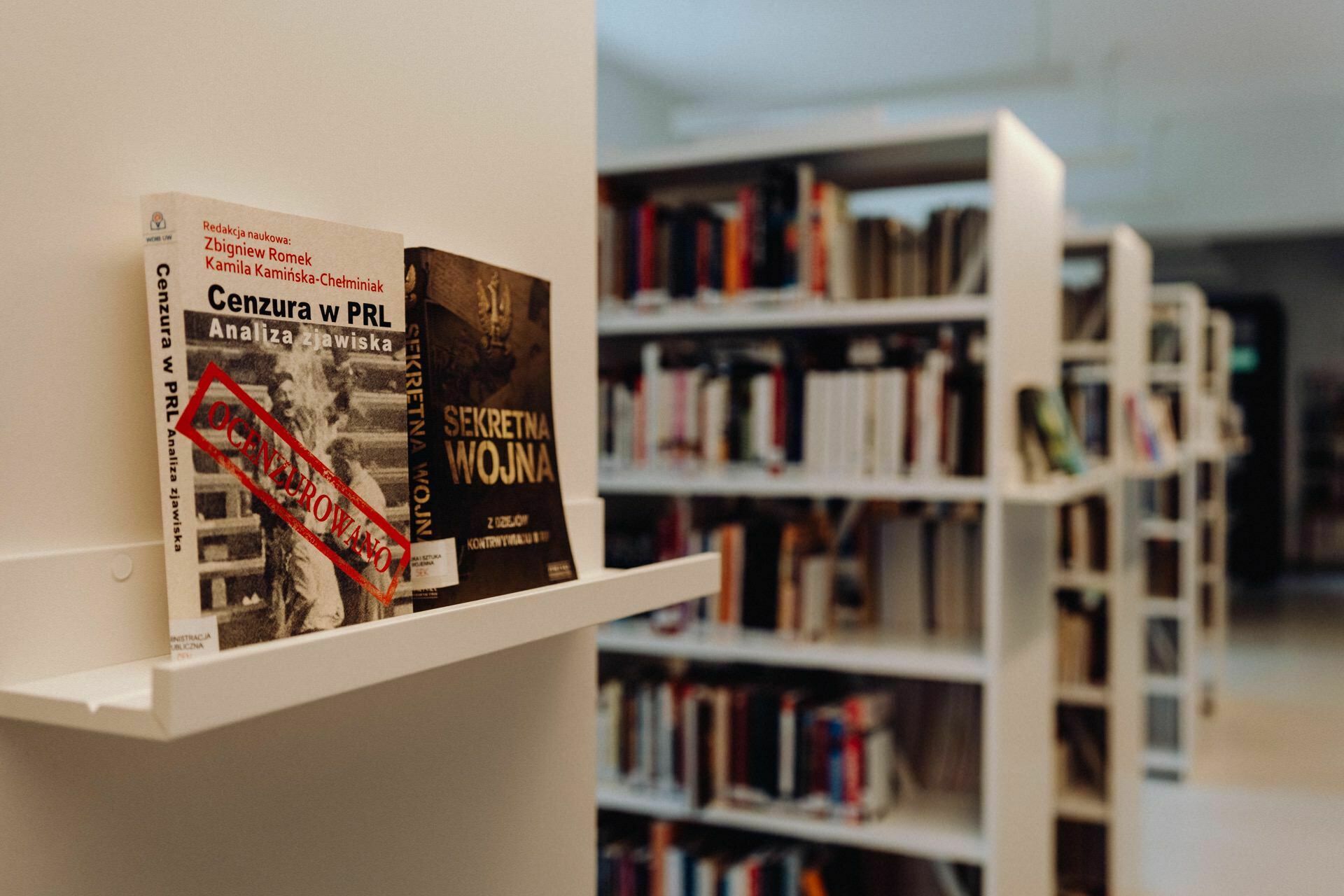 Bookshelves in a library with two books prominently displayed on a white shelf, their titles in Polish. The bookshelf and books are in focus, and the shelves in the background with additional books are slightly blurred, reminiscent of an ingenious composition made by a photographer at an event. 