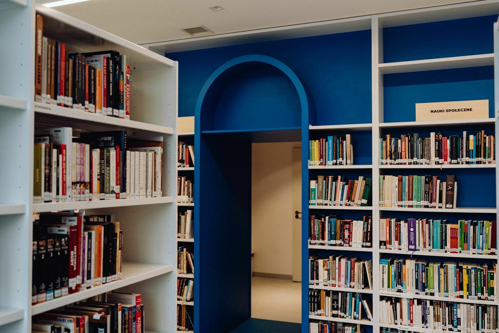 A modern library interior with tall white bookshelves filled with neatly stacked books. In the background is a blue passage leading to another room. On the shelf to the right is a small signboard that reads "Social Sciences." The floor and ceiling are also white, creating the perfect setting for Event Photographer Warsaw.   