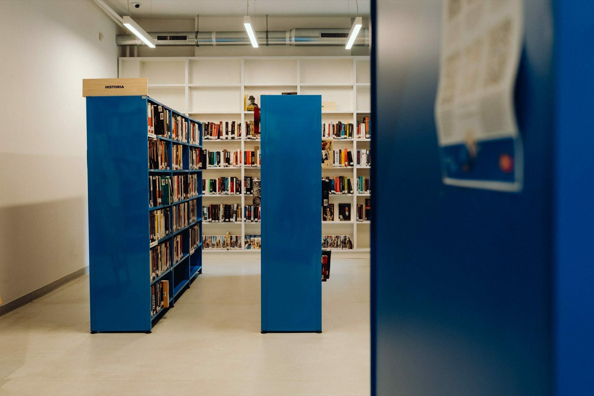 A brightly lit library with rows of shelves filled with books. Two blue bookshelves are visible in the foreground, one with the word "History" written on it. In the background, you can see more shelves filled with books against a white wall, which was perfectly captured in a photo report of the event by event photographer Warsaw. Fluorescent lights hang from the ceiling.   