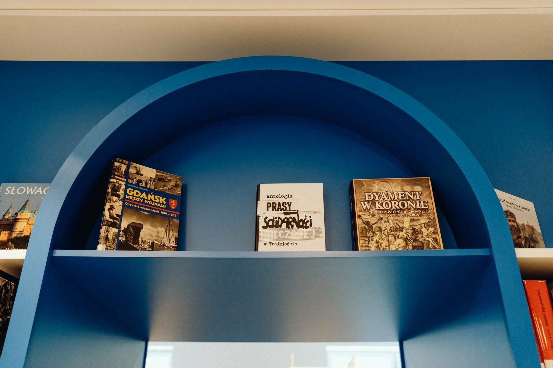 There are four books on a blue arched shelf. From left to right: a book with the coat of arms of Gdansk, a black and white book titled. "Anthology of the Solidarity Fighting Press," and two more books with illustrative covers depicting architectural scenes captured by a photographer from Warsaw.  