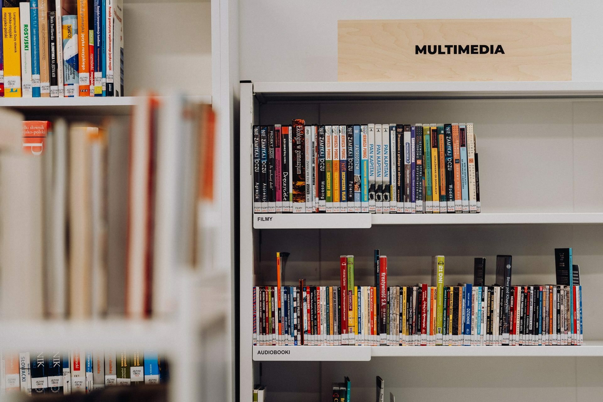 A library shelf labeled "Multimedia" stores DVDs and audiobooks. Rows of various movie and book titles are visible, with labels on the spines indicating genre categories such as "Film" and "Audiobook." To the left, bookshelves are partially visible, capturing the essence like a photo-op of an event.  