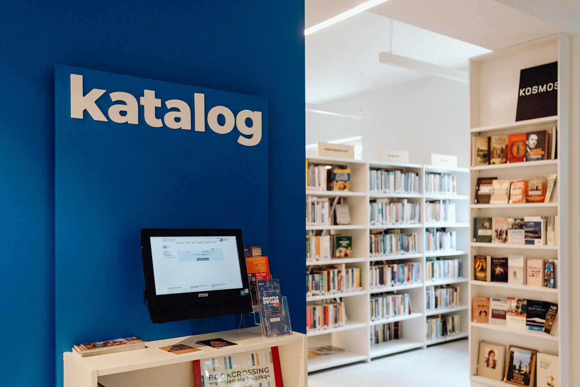 The well-lit interior of the library features a blue kiosk labeled "catalog" with a computer interface for searching the catalog. Book shelves are stocked with a variety of books, and a smaller display shelf with the word "Cosmos" will hold more books and magazines, perfect for capturing in a photo essay of the event. 