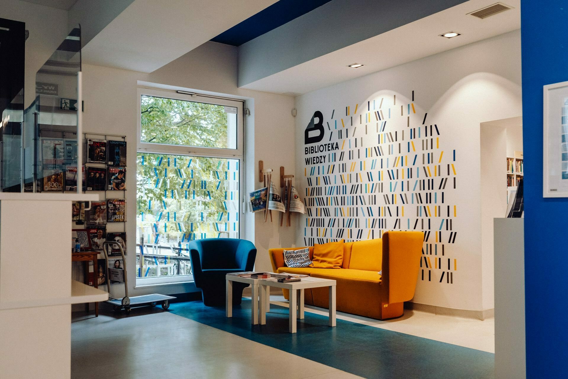 A cozy library corner with modern decor, perfect for quiet reading or photo-shopping at an event. It includes a bright yellow sofa, a small white table and a mustard-colored armchair. The wall behind them is decorated with vertical lines and a "LIBRARY" sign. Large windows let in natural light.   