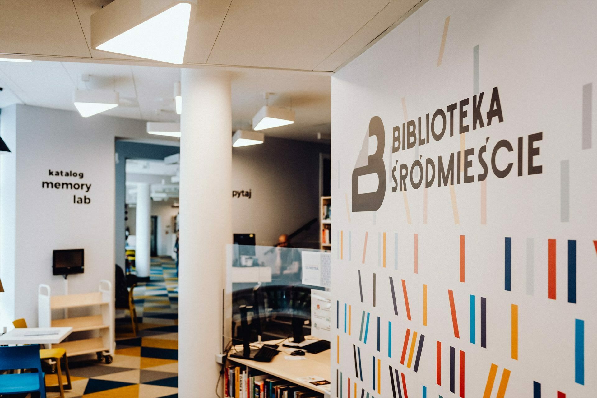 A modern library with colorful geometric patterns on the walls, captured by a photographer for an event from Warsaw. The signs in Polish read: "Downtown Library," "catalog," "memory lab" and "ask." Bookshelves and work areas are visible in the background, with overhead lighting and a white column in the foreground.  