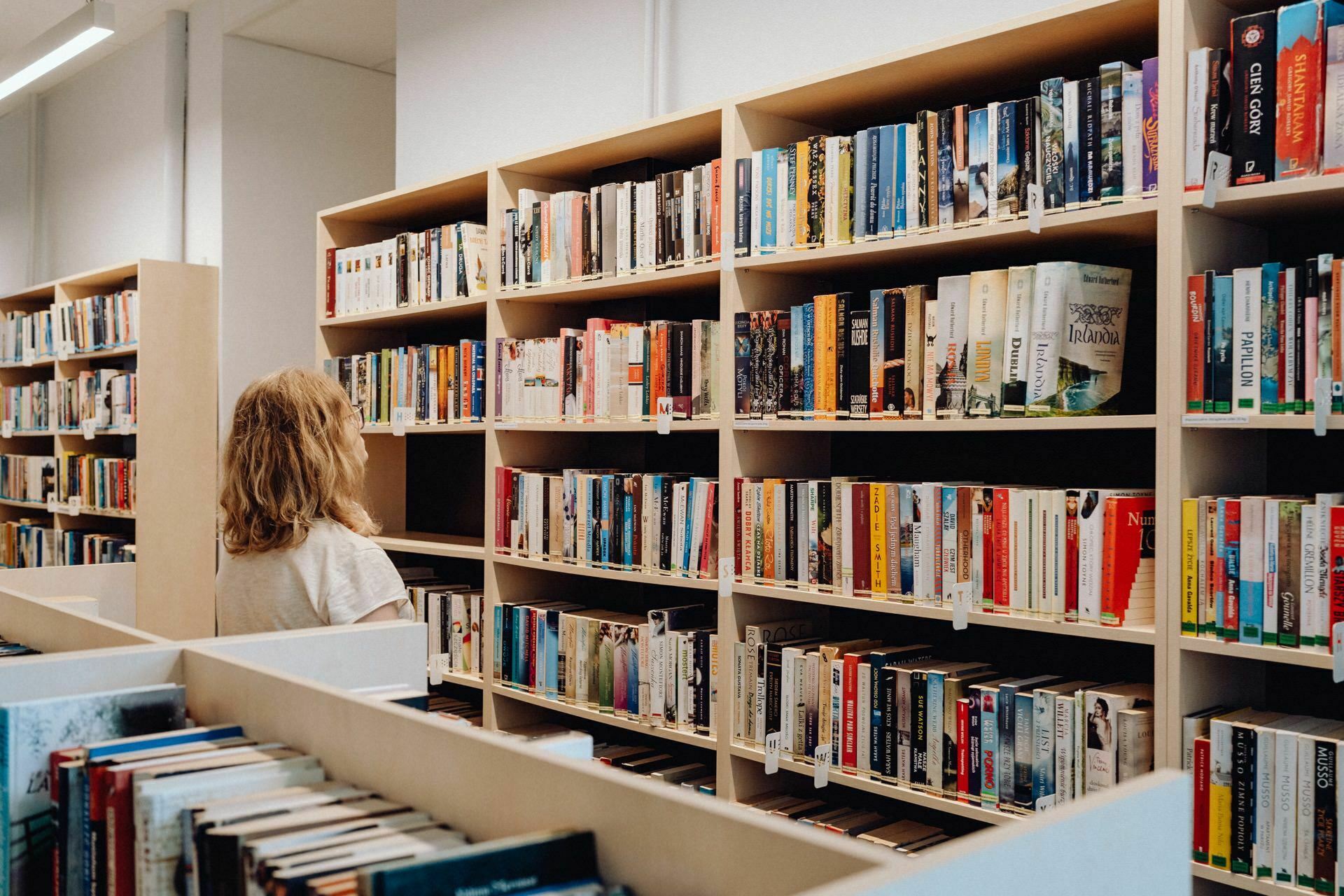 A person with shoulder-length blonde hair stands in a library in front of tall, beige bookshelves filled with various books. The person is browsing the bookshelves, presumably looking for a book. The surroundings are clean and orderly, reminiscent of an artful photograph of an event taken by a talented photographer from Warsaw.  