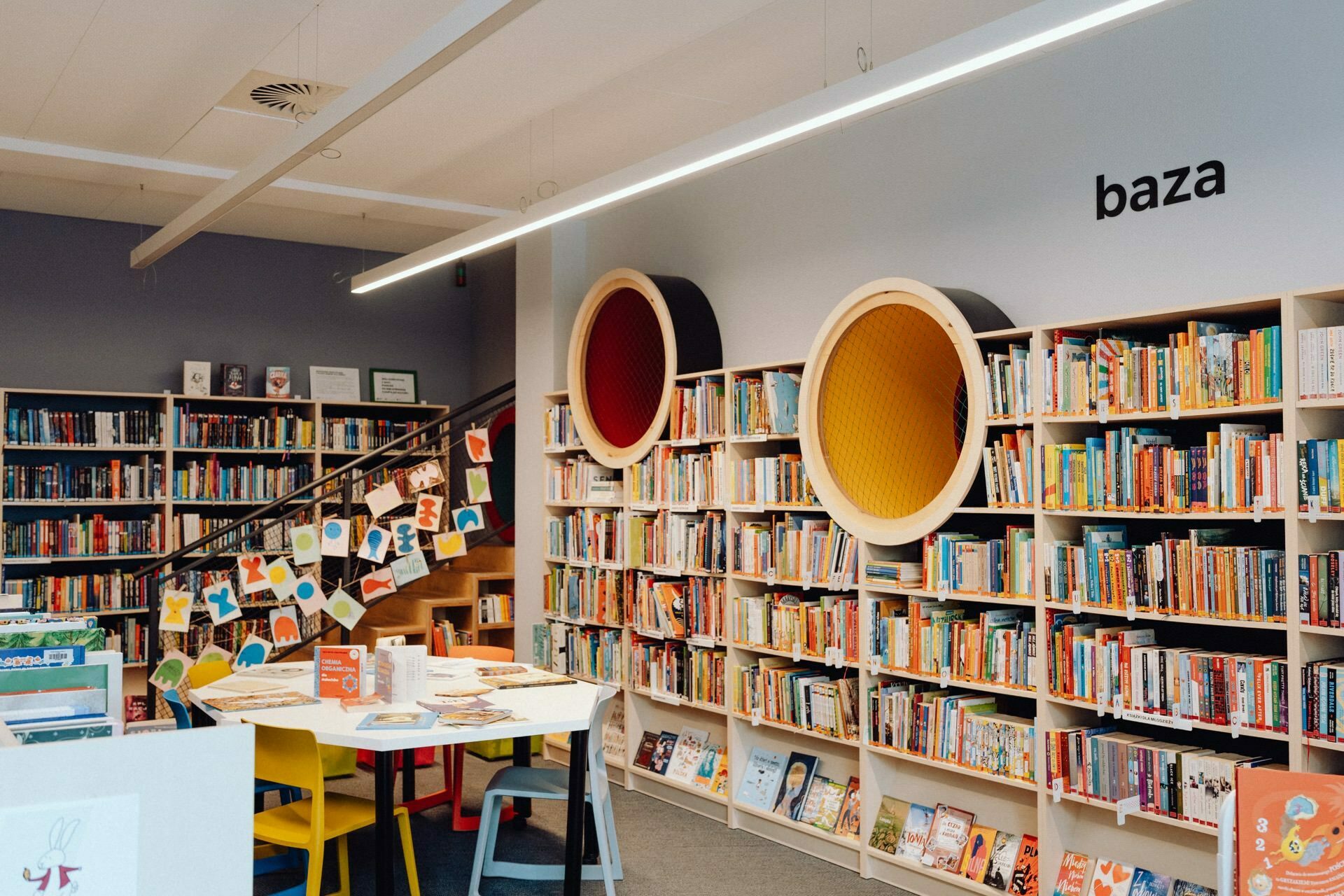 The photo shows a bright and colorful library interior with shelves full of books. There is a seating area with tables and chairs, a display of books and artwork, and two round, recessed, padded seats built into the bookcases. A sign on the wall reads "base," which, like a well-planned photo essay of an event, captures the essence of the event.  