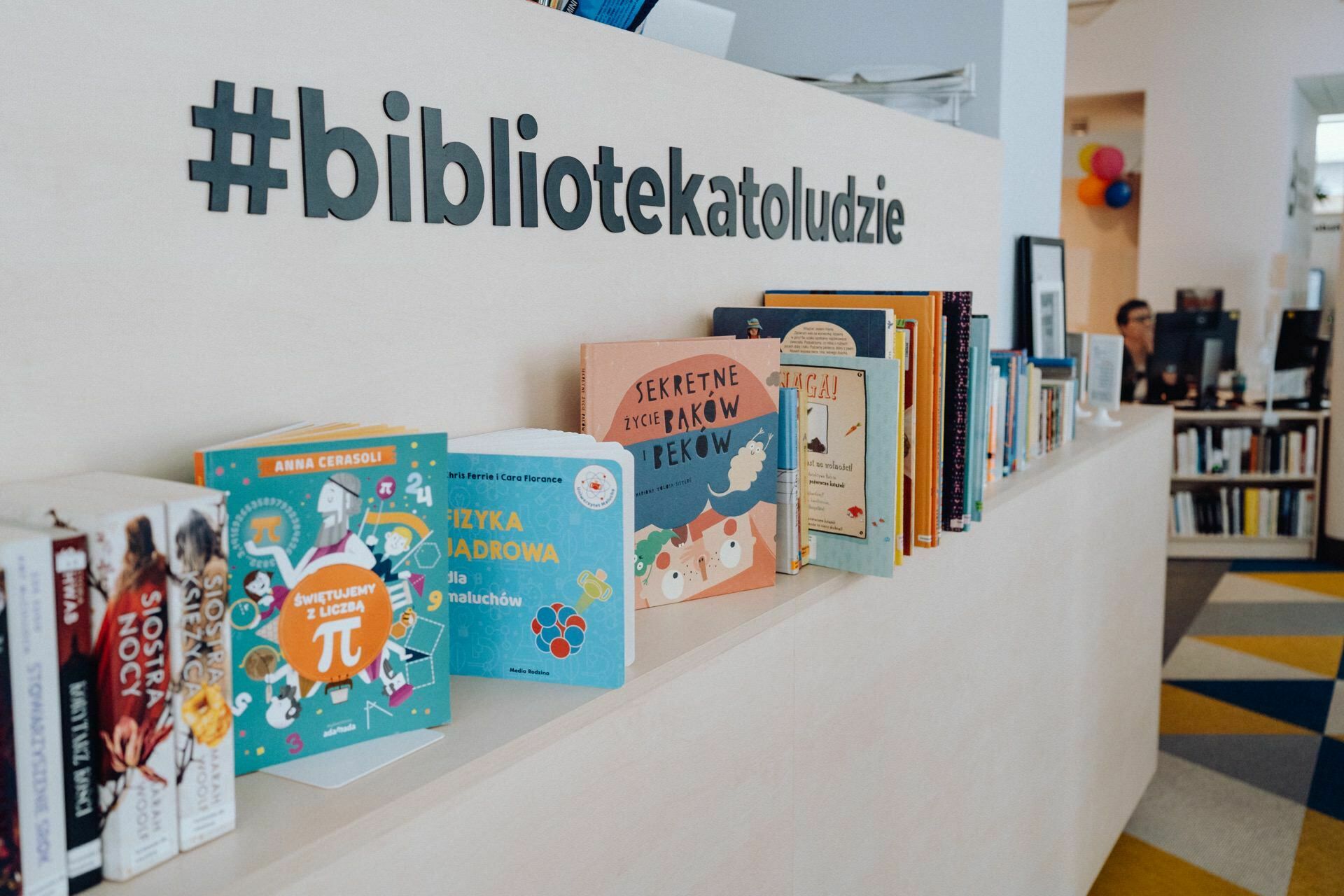 A row of colorful children's books is lined up on a shelf under the hashtag "#bibliotekatoludz". In the background, a Warsaw photographer sits at a desk with books on either side and colorful balloons on the wall. The floor has a geometric pattern.  