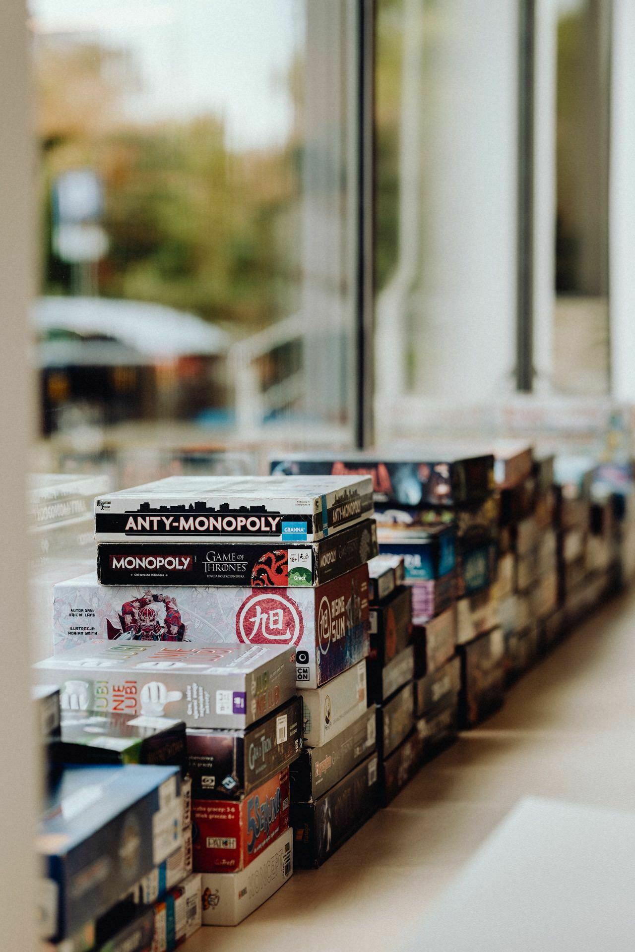 Stacks of various board games are stacked under a large window, creating a colorful and diverse assortment. Visible titles include Anti-Monopoly, Monopoly Game of Thrones and other popular board games. The window lets in natural light, perfect for a photographer at the event to capture a snapshot of the event.  