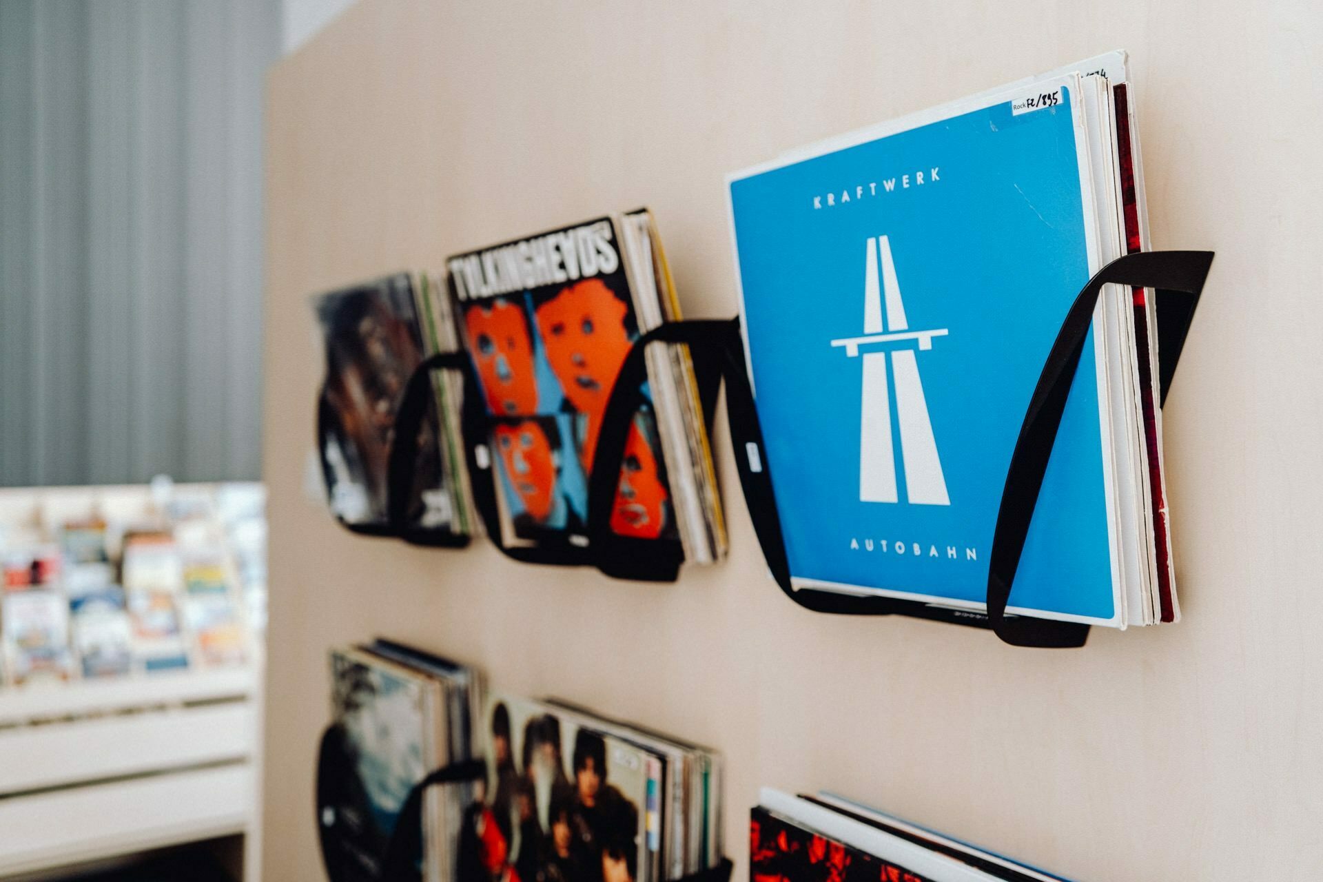 A display of vinyl records stored vertically in wall mounts. Visible covers include an album with a stylized blue background and white artwork by Kraftwerk entitled "Autobahn." In the background, more records and shelves of various items create a scene reminiscent of a photo event.  